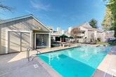 Located at 1420 Lake Blvd in Davis, California, this single-level community outdoor amenity area offers a well-appointed recreational space centered around a generously sized swimming pool with crystal-clear blue water. The pool is complemented by a separate circular spa/hot tub, both finished with classic tiled edges and equipped with stainless steel handrails for easy access. The surrounding concrete deck provides ample room for lounging, with chaise lounge chairs positioned poolside and a stone patio table with umbrella offering a shaded seating option. A wood-sided pool house with sliding glass doors sits adjacent to the pool, providing convenient access to additional facilities. The entire area is enclosed by a wooden privacy fence, creating a secure and tranquil atmosphere. Towering mature trees line the perimeter, offering natural shade and a lush, green backdrop that enhances the overall ambiance. The broader complex features multi-story residential buildings with gabled rooflines, chimneys, and balconies visible in the background, along with a pergola and additional outdoor seating areas. Under the bright California sky, this outdoor space delivers a resort-like setting ideal for relaxation and recreation.