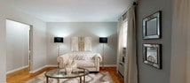 Located at 1076 Stuyvesant Ave in Irvington, NJ, this single-floor residence offers a thoughtfully designed layout with a refined aesthetic throughout. The living room serves as the heart of the home, showcasing classic parquet hardwood flooring, soft neutral-toned walls, and large windows dressed with floor-length drapes that invite abundant natural light. Elegant furnishing choices, including a tufted chesterfield sofa and glass-topped coffee table, enhance the room's sophisticated character. A pass-through window connects the living area to the kitchen, creating an open, airy feel while maintaining distinct zones. The kitchen features light wood cabinetry, granite-style countertops, a stainless steel sink, and white appliances, complemented by clean tile flooring and a window overlooking the outdoors. A secondary room — ideal as a flex space or den — continues the parquet flooring and benefits from generous natural light through patterned curtains. The primary bedroom presents a serene retreat with blue-grey accent walls, a ceiling fan, parquet floors, and ample window light filtered through dark drapes. Throughout the residence, a cohesive neutral and grey palette, decorative accents, and consistent hardwood flooring tie each space together into a warm and polished whole.