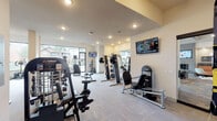 This fitness center in Conroe, Texas offers a comprehensive workout facility thoughtfully organized into distinct training zones across a single floor. Large floor-to-ceiling windows flood the cardio area with natural light and provide views of an outdoor pool and palm-lined surroundings, creating an inviting atmosphere for exercise. The cardio zone is equipped with treadmalls, StairMaster steppers, and elliptical machines, each positioned to take advantage of the outdoor views and wall-mounted flat-screen TVs. A dedicated strength training room features an array of selectorized weight machines, barbell racks with weight plates, and a rack of kettlebells, all set against full-length mirrors and illuminated by track lighting. A functional training area includes TRX suspension trainers, stability balls, and a dumbbell rack, offering versatile options for dynamic workouts. Throughout the space, durable patterned carpet tile flooring provides comfort underfoot, while recessed lighting and a neutral warm color palette maintain a clean, modern aesthetic. Additional amenities include sanitation stations, waste bins, a whiteboard, and an electrical panel area, ensuring the facility is both practical and user-friendly. The overall design balances functionality with a welcoming environment suited for a wide range of fitness routines.