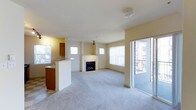 This single-floor residence offers a clean, open layout designed for comfortable everyday living. The living area is generously sized, featuring plush wall-to-wall carpeting that adds warmth underfoot. A corner fireplace serves as a focal point of the main living space, framed by a crisp white mantle that contrasts elegantly against the soft, neutral-toned walls. Natural light flows freely throughout the room via multiple windows fitted with vertical blinds, as well as a full-length sliding glass door that opens to an outdoor balcony, offering views of the surrounding area and further enhancing the sense of openness. The kitchen is seamlessly connected to the living area through a raised breakfast bar, creating an inviting, social layout. Rich wood cabinetry and warm countertops give the kitchen a grounded, functional aesthetic, while a nearby window ensures it remains bright and airy. The overall ambiance of the space is light, neutral, and welcoming, with a consistent color palette of whites and soft creams throughout. The thoughtful flow between the kitchen and living areas makes this a practical and pleasant space for both everyday routines and entertaining.