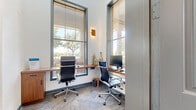 This single-floor business center is thoughtfully designed to accommodate a range of professional needs within a compact footprint. The entrance opens into a stylish lobby featuring warm hardwood-style flooring, soaring ceilings, and an exposed white brick accent wall. A comfortable seating area with a burgundy leather banquette, gray lounge chairs, and a woven ottoman provides a welcoming reception space, while large floor-to-ceiling windows flood the area with natural light and offer views of surrounding greenery.

Adjacent to the lobby, a dedicated workstation area features built-in walnut desks running along two walls beneath tall windows, paired with ergonomic high-back chairs. Natural light pours in through the oversized windows, creating an energizing and productive environment. Patterned carpet tiles add texture underfoot and contrast the sleek, modern furnishings.

A glass-walled conference room accommodates a solid wood meeting table surrounded by professional seating, enhanced by a striking wood-paneled accent wall with a botanical mural. A window with a roller shade provides controlled natural light. Additional individual workstations with built-in cabinetry complete the space, offering functional areas for focused, private work throughout.