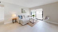 This contemporary two-floor residence is designed with a clean, modern aesthetic throughout. The living area is bright and welcoming, featuring large-format white tile flooring, neutral warm-toned walls, and sliding glass doors that open to a private outdoor terrace, flooding the space with natural light and offering pleasant greenery views. The kitchen is a standout feature, outfitted with rich dark wood cabinetry, granite countertops, a decorative mosaic tile backsplash, and a full suite of stainless steel appliances including a range, microwave, dishwasher, and French door refrigerator — providing both function and visual contrast against the light surroundings. The bedrooms are generously proportioned, each featuring light wood-style flooring, ceiling fans, roller shades, and large sliding glass doors or windows that bring in ample natural light and outdoor views. The primary bathroom is elegantly appointed with a glass-enclosed walk-in shower, a soaking tub, white mosaic tile flooring, and clean white wall finishes. A secondary bathroom offers a tub-shower combination with a glass enclosure, white tile flooring, and a window for natural light. Throughout the space, the consistent use of neutral tones, modern fixtures, and abundant natural light creates a cohesive, serene living environment.