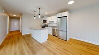 This thoughtfully designed single-floor unit offers a clean, contemporary aesthetic with light hardwood flooring flowing throughout the main living areas. The open-concept kitchen serves as a central highlight, featuring crisp white shaker cabinetry, granite countertops, a central island with pendant lighting overhead, and a full suite of stainless steel appliances including a range, microwave, dishwasher, and refrigerator. The kitchen connects seamlessly to the living area, which benefits from natural light through multiple windows and includes a private exterior door. The bedrooms are well-proportioned, with one featuring a wall-mounted mini-split unit for climate control, baseboard heating, and hardwood floors, while another offers plush carpeting and a bright window. The bathroom is finished with a granite-topped white vanity, a walk-in shower, and matching hardwood-style flooring for a cohesive look. A dedicated laundry area with side-by-side washer and dryer, built-in wire shelving, and additional storage adds everyday convenience. Neutral wall tones and recessed lighting throughout contribute to a bright, welcoming ambiance across the entire unit.
