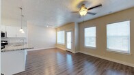 This single-floor space showcases a thoughtfully designed open-concept layout that seamlessly connects the kitchen and living areas. The kitchen is outfitted with crisp white cabinetry, granite countertops, and stainless steel appliances, including a gas range and built-in microwave. A peninsula with an undermount sink serves as both a functional workspace and a natural divider between the kitchen and the adjoining living space. Pendant lighting above the peninsula adds a refined touch, complementing the track lighting installed throughout the main living area. The living room is generously proportioned, featuring warm hardwood-style flooring that runs continuously throughout the space, creating a cohesive and inviting atmosphere. A ceiling fan with an integrated light fixture provides both comfort and additional illumination. Large windows fitted with horizontal blinds allow ample natural light to fill the room, while a glass-paneled door provides access to the exterior. The warm neutral wall tones, textured ceilings, and clean white trim work together to create a bright, welcoming environment that balances contemporary style with everyday functionality.