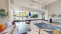 Located in the vibrant Logan Square neighborhood of Chicago, this expansive amenity floor delivers a compelling blend of modern architecture and thoughtfully curated communal spaces. The grand lobby sets an impressive tone with soaring double-height ceilings, floor-to-ceiling glass walls, and polished concrete floors that reflect abundant natural light. A branded entry mat anchors the open foyer, which is furnished with contemporary seating and a sleek marble-topped coffee table, creating a welcoming first impression with lush outdoor greenery visible through the glass facade. Multiple hallways connect the various amenity areas, each lined with large windows that maintain a bright, airy atmosphere throughout. The living room is a relaxed yet sophisticated lounge featuring a fireplace, mounted flat-screen TVs, a billiard table, vibrant wall art, and plush teal velvet seating arranged around a marble accent table. The collaborative area offers a long conference-style table with upholstered chairs alongside glass-enclosed private workrooms, ideal for focused work or small meetings. The dedicated fitness center is well-equipped with strength training machines, free weights, resistance equipment, and open floor space for functional training, all finished with durable rubber flooring and mirrored walls.