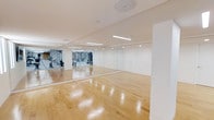This well-appointed fitness facility is designed to support a comprehensive workout experience within a clean, modern environment. The space is anchored by a bright, open-plan exercise studio featuring gleaming polished hardwood floors that reflect the recessed and strip lighting above, creating an airy and energizing atmosphere. Floor-to-ceiling mirrors line one wall of the studio, visually expanding the space while allowing users to monitor their form during workouts. The studio's minimalist design is defined by crisp white walls and a smooth white ceiling, lending a sleek and professional aesthetic throughout. Adjacent to the open studio, the gym area is visible through the mirrored wall and is outfitted with a variety of strength and fitness equipment, providing a fully functional training environment. A flush-fitted white door offers seamless access between areas, maintaining the space's clean and uncluttered visual flow. The recessed lighting and linear strip fixtures ensure even, consistent illumination across the entire floor. Whether used for group fitness classes, personal training, yoga, or free-weight workouts, this single-level facility is thoughtfully designed to accommodate a wide range of physical activities in a polished and professional setting.