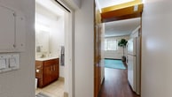 This efficiently designed single-floor unit offers a well-organized layout that maximizes every square foot of its compact footprint. The open-concept living area serves as the heart of the space, featuring warm hardwood flooring, a textured ceiling, and a window that allows natural light to brighten the room. The living area flows seamlessly into a functional kitchen, which is outfitted with rich, warm-toned wood cabinetry, a white refrigerator, a range, and a countertop that doubles as a breakfast bar with seating — an ideal setup for casual dining. The integrated layout makes the space feel cohesive and practical for everyday living. The bathroom is thoughtfully appointed with tile flooring, a warm wood vanity with ample cabinet storage, a white countertop, a mirror, and a walk-in shower with a glass enclosure. A washer and dryer unit is also conveniently accessible within the space, adding a practical amenity. Throughout the unit, the consistent use of warm wood tones in the cabinetry and flooring creates a cohesive and inviting aesthetic. The overall ambiance is clean, functional, and well-suited for those seeking an efficient, self-contained living space.