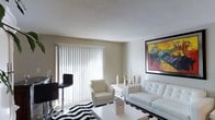 This single-floor space showcases a clean, contemporary living area designed with a minimalist aesthetic. The living room is the focal point of the space, featuring neutral-toned walls that serve as a versatile backdrop for modern décor. A large window dressed with white vertical blinds allows natural light to filter into the room, creating a bright and airy atmosphere.

The living area is furnished with a sleek white leather sofa and a matching armchair, both contributing to the room's cohesive, modern style. A mirrored console table adds a touch of glam, adorned with gold decorative accents that complement the overall color palette. A bold, large-scale abstract painting in warm tones of yellow, red, and green serves as a striking focal point on one wall, injecting vibrant color into the otherwise neutral space.

The flooring is carpeted and partially covered by a bold black-and-white chevron area rug, adding graphic contrast and visual interest. The textured ceiling adds subtle architectural detail throughout. The overall ambiance is polished and stylish, with a careful balance of light tones and bold accents.