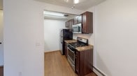 This single-floor unit offers a practical and well-organized layout across several distinct areas. The living space is finished with rich hardwood-style flooring and features a large sliding glass door dressed with vertical blinds, allowing natural light to fill the room. A wall-mounted air conditioning unit and baseboard heating ensure year-round comfort.

The kitchen is efficiently designed with dark espresso cabinetry, granite-style countertops, and a full suite of stainless steel appliances, including a gas range, over-the-range microwave, and refrigerator. Light hardwood-style flooring and overhead lighting complete the space.

The bedroom is a calm, neutrally toned room with plush carpeting and a large window fitted with vertical blinds that invite natural light. Baseboard heating runs along the perimeter, and the clean white walls provide a versatile backdrop.

The bathroom is finished with classic cream-toned tile surrounding a full bathtub and shower combination. A wall-mounted pedestal sink sits beneath a well-lit vanity mirror, with a medicine cabinet and open wood shelving providing storage. Small mosaic floor tiles add a traditional touch to the overall clean and functional design.