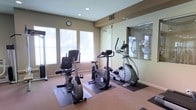 This fitness center is designed to support a well-rounded cardio workout in a clean, functional environment. The space is outfitted with a selection of cardio equipment, including a recumbent bike, an upright stationary bike, and an elliptical trainer, each placed on protective floor mats for safety and stability. A large mirrored wall panel runs along one side of the room, visually expanding the space and allowing users to monitor their form during exercise. The neutral-toned walls and carpeted flooring contribute to a calm, focused atmosphere. Large windows along the exterior wall flood the room with natural light, creating an open and inviting feel while offering views of surrounding greenery. Recessed ceiling lights provide additional illumination for evening or low-light use. A wall clock keeps exercisers on schedule, adding a practical touch to the room's layout. The glass-paneled partition visible on one side suggests a connection to an adjacent space, such as a studio or additional fitness area. Overall, the fitness center offers a straightforward, comfortable environment well-suited for daily cardio training.
