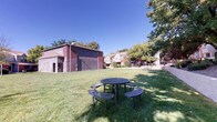 Located at 5801 Eubank Blvd NE in Albuquerque, New Mexico, this single-level outdoor common area serves as a central gathering space within a residential community. The grounds are defined by well-maintained, expansive green lawns that provide an open and inviting atmosphere beneath the characteristically clear blue skies of the Southwest.

The space features multiple outdoor picnic tables with attached bench seating, positioned across the lawn to encourage social gatherings and relaxation. Mature deciduous and evergreen trees are distributed throughout the grounds, offering natural shade and visual landscaping interest. Raised concrete planters and low retaining walls divide portions of the grounds, adding structured definition to the open space.

The surrounding residential buildings are constructed with classic brick and stucco exteriors in warm earth tones, contributing to a cohesive architectural character. Waste receptacles and utility fixtures are conveniently placed throughout the grounds to support everyday use. The overall layout promotes ease of movement across the property, with clear sightlines between the various outdoor areas.

The combination of lush greenery, functional seating, and the surrounding architectural backdrop creates a welcoming communal environment well-suited for outdoor leisure and neighborly interaction in the heart of Albuquerque.