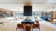 Located at 2025 Fulton Place in Reston, Virginia, this single-floor communal amenity space is thoughtfully designed to foster comfort and community. The centerpiece of the main lounge is a striking suspended double-sided fireplace with a bold dark surround, positioned above a richly finished square hearth base and flanked by carefully arranged seating that encourages both conversation and relaxation. Light hardwood flooring runs throughout the space, complementing warm wood-paneled walls that lend a refined, natural aesthetic. Floor-to-ceiling glass doors and expansive windows flood the interior with abundant natural light while offering direct views of and access to an outdoor courtyard. A built-in library wall, fully stocked with books, serves as both a functional and decorative feature, anchoring a reading and lounge area furnished with contemporary chairs and low tables. A generously sized built-in banquette with leather upholstery runs along one side of a stepped lower-level seating zone, accompanied by modern accent chairs in muted tones and marble-topped side tables. Subtle recessed lighting, cove lighting accents, and warm-toned pendant fixtures enhance the ambiance throughout. Black-framed glass partition doors with frosted panels provide access to additional rooms, while an open staircase with dark brick detailing adds architectural character to the overall design.