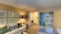 This single-floor residence offers a thoughtfully arranged layout that balances comfort and functionality. The living area serves as the heart of the space, featuring plush carpeted flooring and a warm, ambient lighting scheme that creates an inviting atmosphere. A large white sofa anchors the room, complemented by accent chairs and decorative throw pillows, while framed wall art and a bold blue abstract painting add personality and visual interest to the space.

A textured room divider elegantly separates the living area from the adjoining dining and kitchen zone, maintaining an open and airy feel while defining each functional area. The kitchen is finished with crisp white cabinetry and light-toned countertops, offering a clean and classic aesthetic. A compact dining area with a dark bistro-style chair provides a casual spot for meals just off the kitchen.

The textured ceiling adds a subtle architectural detail throughout, and the warm, golden lighting enhances the cozy ambiance across all areas. The overall design blends neutral tones with carefully chosen accent pieces, resulting in a space that feels both livable and stylishly curated — ideal for comfortable everyday living.