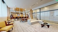 This upscale amenity lounge is designed with a refined, contemporary aesthetic that seamlessly blends multiple social and recreational zones within a single open-plan floor. The space is anchored by a herringbone-patterned light hardwood floor that extends throughout, contrasting elegantly with warm wood-paneled walls and a recessed wood-clad ceiling detail in the billiards area.

The games zone features a sleek white billiards table positioned beneath a sculptural linear pendant light, with a wall-mounted television and a built-in wet bar with dark wood cabinetry nearby. A glazed partition wall with floor-to-ceiling black-framed glass panels creates a transparent boundary, maintaining visual openness while defining separate areas.

The lounge zone is furnished with a large sectional sofa in neutral tones, mid-century-inspired wooden dining chairs around a round table, and mustard-yellow accent armchairs arranged on a patterned area rug. A full media wall with open white shelving and a wall-mounted television anchors this seating area. Sheer floor-to-ceiling curtains diffuse natural light along the perimeter, contributing to the bright, airy ambiance throughout the space.