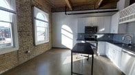 This single-floor space showcases a compelling blend of industrial heritage and contemporary design. The open layout is anchored by striking exposed brick walls that add warmth and texture, while overhead, original timber beams and visible ductwork reinforce the loft's raw, industrial aesthetic. Polished concrete floors run throughout, providing a sleek, low-maintenance surface that complements the space's character.

A large arched window dominates one wall, flooding the interior with abundant natural light and offering views of the surrounding urban environment. The sunlight casts dramatic arched reflections across the walls, enhancing the open, airy feel of the space.

The kitchen area is outfitted with white cabinetry, dark countertops, and stainless steel appliances including a range and over-the-range microwave. A dark-topped island on casters provides flexible workspace and can be repositioned as needed. The contrast between the crisp white cabinetry and the rugged brick and timber elements creates a balanced interplay between modern utility and historic charm, making this space both functional and visually distinctive.