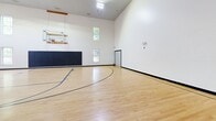 Located at 870 Hartford Tpke in Shrewsbury, MA, this expansive amenity facility offers a comprehensive range of spaces designed to support both active recreation and social gatherings. The centerpiece is a soaring full-size indoor basketball court featuring polished hardwood floors with regulation court markings, padded wall protection, mounted hoops, and dramatically high ceilings that create an impressive sense of volume and openness. Large windows allow natural light to fill the space while maintaining a clean, bright atmosphere. Adjacent to the court, the well-appointed fitness center is divided into dedicated zones for cardio and strength training. The cardio area houses treadmills, ellipticals, stair climbers, and stationary bikes, while the strength zone offers free weights, cable machines, and wall-to-wall mirrors. Durable speckled flooring and overhead lighting complete the functional layout. The social hub of the facility is a light-filled lounge and dining area featuring hardwood floors, sheer-draped windows with lush outdoor views, and comfortable seating. The adjoining kitchen is fully equipped with stainless steel appliances, gray shaker cabinetry, granite countertops, a decorative tin-paneled island with bar seating, and tile flooring — making it ideal for community events and gatherings.