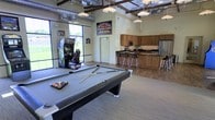 This single-floor recreational space is designed to entertain and bring people together. The open-concept layout is anchored by a full-size pool table positioned at the center of the room, inviting friendly competition. Adjacent to the billiards area, an arcade racing game adds a lively, playful atmosphere to the space. Sports memorabilia, including a prominent Chicago Cubs sign, lends the room a spirited, fan-friendly character.

The fully equipped kitchen runs along the back wall and features warm wood cabinetry, dark granite countertops, stainless steel appliances including a refrigerator and microwave, and a tiled backsplash. A breakfast bar with sleek bar stools provides additional seating and a natural gathering point.

The interior design showcases exposed structural beams and ductwork along the ceiling, contributing to an industrial-chic aesthetic balanced by warm, neutral wall tones and light hardwood-style flooring. Pendant lighting hangs throughout, providing warm, even illumination across the space. Large windows along one wall flood the room with natural light, creating an open and welcoming ambiance. This versatile space is well-suited for social gatherings, casual recreation, and community events.
