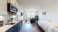 This single-floor studio unit presents a thoughtfully designed open-plan layout that efficiently integrates the kitchen, living, and sleeping areas into one cohesive space. The interior is finished with warm-toned hardwood flooring throughout, crisp white walls, and recessed lighting that creates a bright, clean ambiance.

The kitchen is a standout feature, showcasing a two-tone cabinet design with bold navy lower cabinets paired with white upper cabinets, complemented by a white subway tile backsplash and light quartz countertops. Stainless steel appliances, including a built-in microwave, dishwasher, and refrigerator, provide a complete and modern cooking environment with a single-basin sink and gold-toned faucet.

The open living area flows naturally from the kitchen, offering space for a sofa, coffee table, and dining area. A pair of double-hung windows along the far wall admit natural light and provide views of the surrounding neighborhood. A wall-mounted television adds convenience to the layout.

The sleeping area is seamlessly integrated into the open floor plan. The bathroom features classic small-format hexagonal floor tiles, a walk-in shower with curtain, toilet, and a white vanity with ample cabinetry.