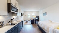 This single-floor studio unit presents a thoughtfully designed open-plan layout that efficiently integrates the kitchen, living, and sleeping areas into one cohesive space. The interior is finished with warm-toned hardwood flooring throughout, crisp white walls, and recessed lighting that creates a bright, clean ambiance.

The kitchen is a standout feature, showcasing a two-tone cabinet design with bold navy lower cabinets paired with white upper cabinets, complemented by a white subway tile backsplash and light quartz countertops. Stainless steel appliances, including a built-in microwave, dishwasher, and refrigerator, provide a complete and modern cooking environment with a single-basin sink and gold-toned faucet.

The open living area flows naturally from the kitchen, offering space for a sofa, coffee table, and dining area. A pair of double-hung windows along the far wall admit natural light and provide views of the surrounding neighborhood. A wall-mounted television adds convenience to the layout.

The sleeping area is seamlessly integrated into the open floor plan. The bathroom features classic small-format hexagonal floor tiles, a walk-in shower with curtain, toilet, and a white vanity with ample cabinetry.