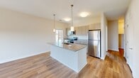 Located in downtown Concord, NC, this well-appointed 2-bedroom, 2-bathroom residence offers a thoughtfully designed open-concept layout. The combined kitchen and living area serves as the heart of the home, featuring warm wood-look flooring throughout. The kitchen is a standout, equipped with a large central island topped with granite countertops, a undermount sink, stainless steel appliances including a refrigerator, range, and microwave, and light wood cabinetry accented by stylish pendant lighting. The living area is bright and airy, with French doors that open to a private balcony overlooking the surrounding streetscape, filling the space with natural light. Both bedrooms are carpeted for comfort and include ceiling fans, with large windows offering views of the Concord skyline. The primary bedroom is generously sized, while the second bedroom provides versatile use. Both bathrooms are finished with granite-topped vanities and light wood cabinetry; one features a walk-in glass shower with white tile surround, while the other offers a full bathroom setup. Neutral tones and clean lines throughout create a cohesive, modern aesthetic suited for comfortable urban living.