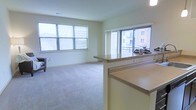 This single-floor residence offers a clean, open-concept layout that connects the kitchen and living areas in a cohesive, functional design. The kitchen features a peninsula with granite-style countertops, providing both additional prep space and a natural divider between the cooking and living zones. Complementary laminate counters extend along the kitchen workspace, offering ample surface area for everyday use.

The living area is finished with plush wall-to-wall carpeting in a neutral tone, creating a comfortable and inviting atmosphere. Large windows line the exterior wall, flooding the space with abundant natural light throughout the day. These windows also frame views of the surrounding neighborhood and an adjacent balcony or outdoor walkway, enhancing the sense of openness and connection to the outdoors.

The interior palette is kept light and neutral throughout, with soft cream-toned walls that complement the natural brightness of the space. The overall ambiance is airy and welcoming, well-suited for both everyday living and entertaining. The thoughtful flow between the kitchen and living areas makes the space feel expansive and practical, while the generous window placement ensures a consistently bright and pleasant environment.