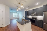 This single-floor residence showcases a thoughtfully designed open-concept layout that seamlessly connects the kitchen and living areas. The kitchen serves as the centerpiece of the space, featuring dark slate-toned shaker-style cabinetry accented with brushed gold hardware. A substantial center island with a dark granite countertop and an integrated sink is illuminated by a trio of elegant brass pendant lights, creating a striking focal point. A herringbone-patterned tile backsplash adds textural detail behind the range, while stainless steel appliances, including a refrigerator, electric range, and over-the-range microwave, provide a polished, contemporary finish.

Rich hardwood-style flooring extends throughout the main living area, which is brightened by a window and ceiling fan. The open layout provides a generous, versatile living space. The bedroom features plush wall-to-wall carpeting, a ceiling fan, and a window allowing natural light, with direct access to the bathroom and an adjoining room. The bathroom is cleanly appointed with large-format neutral tiles, a soaking tub with brushed gold fixtures, black matte hardware accents, dark granite countertop, and built-in open shelving for convenient storage.