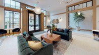 This elegantly designed lobby lounge showcases a refined blend of modern sophistication and warm architectural detailing. The space is defined by soaring ceilings with clerestory windows framed in natural wood trim, flooding the interior with an abundance of natural light. Large black-framed entry doors open to a welcoming street-facing facade, while the generous open floor plan accommodates multiple seating arrangements with ease.

The flooring transitions between classic white penny-tile mosaic and soft area rugs, anchoring distinct zones within the space. A central lounge area is furnished with deep emerald green velvet sofas accented by richly toned throw pillows, centered around a solid wood coffee table topped with artful floral arrangements. A statement globe pendant light hangs overhead, adding a warm, sculptural focal point.

A cluster of hexagonal gold-framed mirrors adorns one wall, reflecting light and adding a contemporary artistic touch, complemented by understated wall sconces. An intimate seating nook with navy velvet chairs and a round bistro table provides a quieter retreat. A staircase with warm wood railings and metal balusters is visible through an interior framed opening, connecting to additional levels. Lush potted plants throughout the space add a fresh, vibrant contrast to the neutral palette.