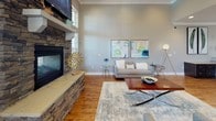 Located on Oak Springs Drive in Greenville, South Carolina, this single-story space presents a well-appointed, open interior designed to accommodate a variety of functions. Warm hardwood flooring flows consistently throughout, unifying each area and contributing to a cohesive, welcoming atmosphere.

The main living and lounge area is anchored by a striking floor-to-ceiling stacked stone fireplace with a mounted television above, complemented by high ceilings that enhance the sense of openness. Natural light enters generously through multiple windows fitted with plantation shutters. Contemporary artwork and decorative wall pieces add character throughout.

An adjacent workspace features a built-in L-shaped desk with dark cabinetry, accommodating multiple workstations with ample counter space — well-suited for administrative or leasing functions.

A dedicated meeting or dining area includes a high-top table with upholstered seating beneath a modern multi-bulb chandelier, with tall windows providing an abundance of natural light and outdoor views.

The kitchen is outfitted with dark cabinetry, granite countertops, stainless steel appliances, and a center island with seating. Elevated ceilings and large windows make this area feel bright and spacious, completing the functional and polished character of the overall space.