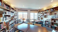 This single-floor library is thoughtfully designed to serve as both a quiet reading retreat and a social gathering space. Floor-to-ceiling built-in wooden bookshelves line the walls, filled with an extensive collection of books and accented by warm wall-mounted lighting that highlights a textured, decorative backdrop. Large windows fitted with roller shades allow abundant natural light to filter in while offering views of the surrounding neighborhood.

The space is organized into distinct activity zones. A central area features rectangular wooden tables with white upholstered chairs, suitable for reading, puzzles, or group activities. A dedicated game table surrounded by tufted blue chairs provides a comfortable setting for board games and puzzles. A separate lounge area with cushioned armchairs offers a more relaxed seating option.

Additional amenities include a water fountain station, a glass-paned private room accessible via a white door, and framed artwork displayed throughout, including a vivid sunset landscape and a linear equestrian sketch. Recessed ceiling lighting complements the natural light, ensuring the space remains bright and welcoming. The carpeted flooring adds warmth and comfort, contributing to the library's inviting and functional ambiance.