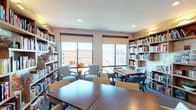 This single-floor library is thoughtfully designed to serve as both a quiet reading retreat and a social gathering space. Floor-to-ceiling built-in wooden bookshelves line the walls, filled with an extensive collection of books and accented by warm wall-mounted lighting that highlights a textured, decorative backdrop. Large windows fitted with roller shades allow abundant natural light to filter in while offering views of the surrounding neighborhood.

The space is organized into distinct activity zones. A central area features rectangular wooden tables with white upholstered chairs, suitable for reading, puzzles, or group activities. A dedicated game table surrounded by tufted blue chairs provides a comfortable setting for board games and puzzles. A separate lounge area with cushioned armchairs offers a more relaxed seating option.

Additional amenities include a water fountain station, a glass-paned private room accessible via a white door, and framed artwork displayed throughout, including a vivid sunset landscape and a linear equestrian sketch. Recessed ceiling lighting complements the natural light, ensuring the space remains bright and welcoming. The carpeted flooring adds warmth and comfort, contributing to the library's inviting and functional ambiance.