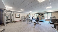 The fitness center at 200 E Burnsville Pkwy in Burnsville, MN is a dedicated exercise facility housed within a single, well-organized room. The space features a drop-panel ceiling with recessed fluorescent lighting, providing consistent, even illumination throughout. Patterned carpet flooring covers the entire area, adding comfort underfoot during workouts.

The cardio zone is anchored along a natural brick accent wall, where large windows offer views of the surrounding outdoor environment, allowing ample natural light to fill the space. This area is equipped with multiple treadmills and an elliptical trainer, accompanied by a wall-mounted flat-screen television for entertainment during cardio sessions.

The strength training area is equally well-appointed, featuring a multi-station cable machine, several selectorized strength machines, and adjustable weight benches. A dedicated free weight station includes a full rack of dumbbells, catering to a range of resistance training needs.

Decorative framed artwork adorns the neutral-toned walls, complemented by a bold navy blue accent wall that adds a modern, energetic feel to the space. The room also includes a windowed entry vestibule, providing a clear transition into the facility. Overall, the fitness center offers a comprehensive and functional environment suitable for a full range of workout routines.
