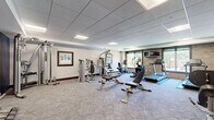 The fitness center at 200 E Burnsville Pkwy in Burnsville, MN is a dedicated exercise facility housed within a single, well-organized room. The space features a drop-panel ceiling with recessed fluorescent lighting, providing consistent, even illumination throughout. Patterned carpet flooring covers the entire area, adding comfort underfoot during workouts.

The cardio zone is anchored along a natural brick accent wall, where large windows offer views of the surrounding outdoor environment, allowing ample natural light to fill the space. This area is equipped with multiple treadmills and an elliptical trainer, accompanied by a wall-mounted flat-screen television for entertainment during cardio sessions.

The strength training area is equally well-appointed, featuring a multi-station cable machine, several selectorized strength machines, and adjustable weight benches. A dedicated free weight station includes a full rack of dumbbells, catering to a range of resistance training needs.

Decorative framed artwork adorns the neutral-toned walls, complemented by a bold navy blue accent wall that adds a modern, energetic feel to the space. The room also includes a windowed entry vestibule, providing a clear transition into the facility. Overall, the fitness center offers a comprehensive and functional environment suitable for a full range of workout routines.