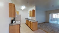 This single-floor space is designed with functionality and comfort in mind. The kitchen serves as a central feature, outfitted with warm oak cabinetry both above and below the countertops, providing ample storage. The gray laminate countertops run the length of the kitchen, offering generous workspace for meal preparation. A double-basin sink is positioned along the lower cabinetry, accompanied by a white dishwasher. White appliances, including a coil-burner electric range and a full-size refrigerator, complete the kitchen setup. The tile flooring in the kitchen offers a clean, easy-to-maintain surface. The kitchen opens directly into an adjacent living area, which transitions to carpeted flooring, creating a clear and practical distinction between the two spaces. A window in the kitchen allows for natural light to filter in, brightening the area. The neutral white and light blue walls throughout contribute to an airy, open atmosphere. Overall, the layout promotes a straightforward, connected flow between the kitchen and living spaces, making the unit practical for everyday living.