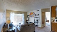 This single-floor space centers around a carpeted living room that serves as the main gathering area. The room is furnished with a neutral-toned sofa and accent chairs arranged around a glass-top coffee table, creating a comfortable seating arrangement. Two lean-style ladder shelves flank a vibrant floral canvas print on the main wall, adding a decorative focal point to the space. Natural light flows into the room through a large sliding glass door dressed with sheer patterned curtains and vertical blinds, offering a view of an outdoor fenced area. The textured popcorn ceiling and neutral wall tones provide a clean backdrop throughout. A hallway visible toward the rear of the living area leads further into the home, suggesting additional rooms beyond. Overhead ventilation is provided via ceiling vents, and a baseboard vent is also visible along the lower wall. The carpeted flooring adds warmth underfoot and runs consistently through the main living area, contributing to the overall cozy and functional ambiance of the space.