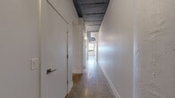 Located at 206 North Ave in Webster, NY, this single-floor unit showcases an industrial-inspired aesthetic throughout its well-organized layout. The main living area is an open, airy space distinguished by polished concrete floors that reflect natural light streaming through a bank of large windows fitted with roller shades. An exposed ceiling with visible ductwork and a stylish ceiling fan add to the loft-like character of the room, lending a contemporary, urban feel. A secondary room features a similar design sensibility with a painted ceiling, ceiling fan, and earthy tile-like flooring, supported by a distinctive rounded structural column that adds architectural interest. Clean white walls throughout create a bright, neutral backdrop adaptable to a variety of uses. The hallway connects the rooms efficiently, leading to a well-appointed full bathroom. The bathroom is generously sized, finished with polished concrete floors, a white vanity with ample cabinetry, a walk-in shower with a glass door, and a toilet — all presented in a clean, minimalist style. A pass-through counter detail visible in one area hints at a defined functional zone within the open plan. The overall ambiance is modern and versatile, blending industrial elements with clean finishes.