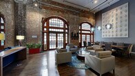 Located at 909 Texas Ave in the heart of downtown Houston, this sophisticated lounge space masterfully blends historic industrial character with contemporary design. The single-floor layout is anchored by soaring ceilings, dramatic exposed brick walls, and a series of grand arched windows framed in rich hardwood, flooding the interior with abundant natural light and offering views of the surrounding urban streetscape.

The main lounge area features warm hardwood flooring throughout and is furnished with modern upholstered seating arrangements centered around a circular glass-top coffee table atop a vibrant area rug. Pendant globe light fixtures hang from exposed ceiling infrastructure, adding a polished industrial aesthetic. A built-in media and bookcase unit with dark walnut cabinetry, open shelving, and integrated storage serves as a functional focal point in the secondary gathering area, complemented by a kitchenette with bar seating, dark cabinetry, and stainless steel appliances.

An outdoor terrace extends the space into the open air, equipped with professional-grade stainless steel grills and lush greenery, set against a striking backdrop of Houston's downtown skyline and neighboring historic architecture. This space offers a refined and versatile environment ideal for work, relaxation, or social gatherings.