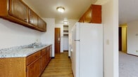 This single-floor unit offers a practical and straightforward layout across several well-defined spaces. The galley-style kitchen is lined with warm wood cabinetry on both upper and lower levels, providing ample storage. A laminate countertop with an integrated sink runs along one wall, complemented by hardwood-style flooring underfoot. White appliances, including a full-size refrigerator, complete the kitchen, which also connects to a closeted storage or utility area at its far end.

The living area is generously proportioned, featuring neutral carpet throughout and a sliding glass door that opens to a balcony, allowing natural light to fill the space and offering views of the surrounding neighborhood. A wall-mounted air conditioning unit and baseboard heating ensure year-round comfort. An additional window further brightens the area.

The bedrooms are carpeted and finished with neutral walls, each equipped with baseboard heating. One bedroom benefits from two windows with views of mature trees, while the other features a single window with neighborhood views. Closet space is accessible from the bedroom area.

The bathroom includes a full tub-shower combination with built-in shelving, a toilet, and a vanity, all finished in clean, light tones with tile flooring.