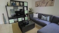 This thoughtfully designed two-floor space offers a comfortable and contemporary living environment. The living area serves as a central gathering point, anchored by a sleek, dark entertainment wall unit with contrasting light green cabinet inserts, providing ample display and storage space. A flat-screen television is seamlessly integrated into the unit, making it a functional focal point of the room. Opposite the entertainment wall, a plush gray sectional sofa with patterned accent cushions creates a welcoming seating area, arranged around a dark, low-profile coffee table that adds a modern edge to the layout. A natural fiber area rug grounds the seating arrangement, lending warmth and texture to the tiled floor. A lush indoor plant adds a refreshing organic element to the corner of the room, complementing the neutral wall tones. Wall art featuring soft floral motifs adds a subtle decorative touch above the sofa. The overall ambiance of the space is modern yet comfortable, with a cohesive color palette of dark tones, grays, and soft greens that ties the design together. The two-floor layout offers additional living and functional areas beyond the main living space, contributing to an overall sense of spaciousness and versatility.