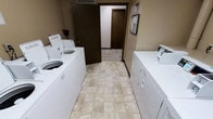 This shared laundry facility is designed for practical, everyday use. The room is laid out with washers and dryers positioned along both side walls, maximizing the usable floor space and allowing multiple users to operate machines simultaneously. The flooring consists of neutral-toned ceramic tiles in a classic grid pattern, offering durability and ease of maintenance. The walls are finished in a warm tan tone, and the room is illuminated with overhead lighting that ensures good visibility throughout the space. A framed bulletin board is mounted near the entrance, likely used for posting building notices or laundry guidelines. The room connects directly to a hallway, which features a dark wood-toned door leading further into the building, providing straightforward access for residents. The layout is straightforward and functional, with enough space between the two rows of appliances to move comfortably while loading and unloading machines. Overall, the laundry room presents a clean, no-frills environment focused entirely on utility and convenience for building occupants.