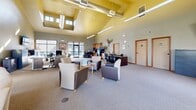 This single-story leasing office is designed to be both functional and welcoming, combining professional workspaces with comfortable lounge areas. The layout is open and generously proportioned, anchored by striking vaulted ceilings with warm yellow tones and exposed ductwork that lend an industrial-modern character to the interior. Pendant and linear light fixtures complement the abundant natural light streaming through large floor-to-ceiling windows and clerestory windows positioned high along the roofline.

The main reception and lounge area features a bold stacked-stone corner fireplace, serving as a focal point alongside a wall-mounted television. Seating arrangements include plush sofas, leather chairs, and accent chairs grouped around coffee tables, creating inviting spaces for prospective residents. A high-top dining table positioned near oversized windows offers additional seating with views of the surrounding community.

The office section includes workstations and a reception desk, providing staff with an organized and accessible area to assist visitors. A refreshment station is also present, adding a hospitable touch. Soft carpeting throughout contributes to a comfortable and professional ambiance, while neutral wall tones and warm wood accents tie the overall aesthetic together cohesively.