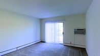 This single-floor space centers around a bright, open main room finished with wall-to-wall carpeting and smooth, light-colored walls that contribute to a clean, neutral aesthetic. A sliding glass door serves as the room's focal point, fitted with vertical blinds that allow for adjustable natural light and outdoor access. The door provides a view of greenery outside, bringing a subtle connection to the outdoors. Baseboard heating runs along the perimeter of the room, ensuring consistent warmth, while a wall-mounted air conditioning unit on the adjacent wall provides climate control year-round. The layout is straightforward and functional, with an open floor plan that offers flexibility in how the space can be arranged and utilized. The pale walls and light-toned ceiling enhance the sense of openness, making the room feel airy despite its modest scale. Overall, the space presents a practical, no-frills interior well-suited for those prioritizing functionality and ease of maintenance.