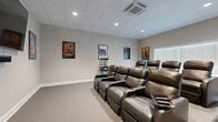 This dedicated home theater room in Longview, Texas is thoughtfully designed to deliver an immersive cinematic experience. The space is configured with two tiered rows of plush, dark leather power recliner seats, each equipped with built-in cup holders, providing comfortable viewing for a group. Seating is arranged on a raised platform at the rear to ensure unobstructed sightlines to the large wall-mounted flat-screen television positioned at the front of the room. A sound bar is mounted directly beneath the screen for enhanced audio performance. The room features a drop ceiling with recessed lighting and a ceiling-mounted HVAC unit, contributing to a well-lit and climate-controlled environment. Neutral-toned walls are adorned with a curated collection of classic movie posters — including Avengers: Infinity War, Rocky, Back to the Future, Jurassic Park, and more — reinforcing the cinematic atmosphere. A popcorn machine stationed along the side wall adds a fun, authentic theater touch. Soft carpeting throughout adds warmth and acoustical benefits. A windowed door and a standard window with blinds allow for natural light when desired. A film reel triptych wall art piece near the rear entry further enhances the movie-themed aesthetic of this well-appointed screening room.