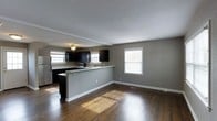 This two-floor home offers a well-designed open-concept layout on the main level, seamlessly connecting the kitchen and living area. The kitchen is outfitted with dark cabinetry that creates a bold contrast against the light grey walls, complemented by a peninsula with a built-in sink that doubles as a breakfast bar. Stainless steel appliances, including a microwave and refrigerator, add a modern touch, while a ceiling fan overhead provides both function and style. Rich, dark hardwood flooring runs throughout the main level, adding warmth and visual depth to the space. Multiple windows dressed with white blinds invite generous natural light, brightening the open living area and highlighting the clean white baseboards and textured ceiling. The neutral grey wall palette creates a versatile backdrop, making the space easy to personalize. The layout promotes an easy flow between cooking and living spaces, ideal for everyday living and entertaining. The second floor offers additional living or sleeping quarters, extending the overall functionality of the home.