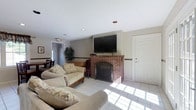 This single-floor space serves as a welcoming communal lounge designed for comfort and casual gatherings. The main living area is anchored by a classic brick fireplace, which serves as the focal point of the room and adds a warm, traditional character to the space. A large wall-mounted flat-screen television is positioned above the fireplace, making it a functional hub for entertainment. Plush, neutral-toned sofas are arranged to encourage conversation and relaxation, complemented by light tile flooring that brightens the room. Natural light streams in through the windows, enhancing the airy feel of the space. A textured ceiling with recessed lighting provides even illumination throughout. A decorative wooden chair rail runs along the walls, adding a subtle architectural detail. Adjacent to the lounge, a dining area is visible, furnished with a table and chairs, extending the utility of the space for meals or group activities. A large potted plant adds a touch of greenery, while an exit-marked doorway ensures easy access and circulation. The overall ambiance is casual, inviting, and well-suited for shared use.