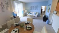 This single-floor residence offers an inviting open-concept layout that seamlessly connects the dining and living areas, creating a cohesive and functional living space. The interior is styled in a calming blue-and-white color palette, with soft blue accent walls serving as a soothing backdrop throughout the main living area.

The dining area is anchored by a warm wood round dining table surrounded by cream-upholstered chairs, set with glassware and decorative dishware for an elegant yet welcoming feel. The space flows naturally into the living area, which features plush, light-toned wall-to-wall carpeting that adds warmth and comfort underfoot.

The living room is furnished with a white sofa accented by blue and green throw pillows, complemented by an oval coffee table and coordinating side tables with table lamps. A gallery wall of framed botanical prints adds a decorative focal point above the sofa. A large sliding glass door and adjacent windows flood the space with abundant natural light, brightening the entire open area and offering a view to an outdoor space beyond.

A doorway visible from the living area suggests additional rooms, hinting at a well-connected floor plan. The overall ambiance is clean, airy, and thoughtfully curated, making the space feel both livable and stylishly appointed.