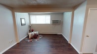 This single-floor space presents a clean and straightforward layout designed for comfortable everyday living. The main living area is grounded by rich, dark hardwood-style flooring that contrasts neatly with the neutral, light-toned walls, creating a warm and inviting atmosphere. A textured ceiling adds a classic finishing touch overhead. Natural light filters in through a horizontal window fitted with white blinds, brightening the space and providing a connection to the outdoors. Climate control is handled by a wall-mounted HVAC unit positioned beneath the window, complemented by a baseboard heater along the exterior wall, ensuring year-round comfort. A recessed overhead soffit detail frames a portion of the room, adding architectural interest and defining distinct zones within the open layout. A framed piece of wall art and a floor lamp add personal touches to the living area, where a small round accent rug with decorative pillows creates a relaxed seating nook. The space offers an efficient and adaptable floor plan, well-suited for a variety of uses while maintaining a tidy, functional character throughout.