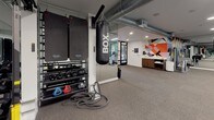 This contemporary fitness center is designed to support a full range of workout routines in a sleek, modern environment. The space is laid out across a single floor with distinct functional zones, each outfitted with purpose-built equipment. The cardio area features treadmills, elliptical machines, stationary bikes, and rowing machines, positioned along floor-to-ceiling windows that frame lush outdoor greenery, flooding the space with natural light. A large flat-screen television and wall-length mirrors enhance the open, energetic atmosphere. The strength and functional training zone is equipped with cable machines, weight racks, kettlebells, medicine balls, and battle ropes, providing versatile options for resistance and conditioning work. A dedicated boxing station, complete with a heavy punching bag and wall-mounted boxing pads, adds a high-intensity training dimension. Exposed industrial-style ductwork runs along the dark ceiling, lending the space a contemporary, urban aesthetic. A bold, colorful geometric mural serves as a vibrant focal point near the entrance area, where a service counter and storage cubbies are conveniently located. The durable rubber flooring throughout ensures safety and comfort during all types of training activities.