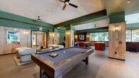 This single-floor amenity space is thoughtfully designed to serve as a multifunctional social hub, blending recreational, dining, and lounge functions within a cohesive, contemporary aesthetic. The interior is anchored by bold sage and forest green walls that contrast warmly against reclaimed wood-clad columns and wall paneling, creating a layered, inviting atmosphere throughout.

At the heart of the space sits a full-sized billiards table with a built-in cue storage column nearby, offering a natural focal point for the recreational zone. Adjacent to it, a comfortable lounge area features curved, bouclé-upholstered chairs arranged around a low coffee table, positioned in front of a dark-tiled bar counter lined with leather barstools and dual wall-mounted flat-screen televisions — ideal for watching sports or casual gatherings.

The kitchenette area is highlighted by a dark tile-clad island, warm wood lower cabinets, and a woven pendant light fixture. A generous bank of windows floods this zone with natural light while framing views of mature trees outside. A casual dining nook nearby features marble-topped tables, mixed seating, and colorful artwork, adding a vibrant, creative energy. Ceiling fans and recessed lighting ensure year-round comfort throughout the space.