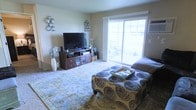 This single-floor space centers around a comfortable living room designed for both relaxation and functionality. The room is finished with smooth, neutral-toned walls that create a clean and inviting backdrop. A large sliding glass door serves as a prominent architectural feature, flooding the space with abundant natural light and providing direct access to an outdoor patio area, where outdoor seating is visible. A wall-mounted air conditioning unit is positioned near the sliding door, ensuring year-round comfort.

The living room is generously proportioned, accommodating a dark wood media console housing a flat-screen television and media components, alongside a patterned upholstered ottoman that doubles as a central coffee table. An ornate area rug with intricate detailing anchors the seating arrangement and adds warmth and texture to the space. Decorative metallic wall art pieces and a tall white floor vase with floral arrangements contribute character and style to the room. The overall ambiance is casual yet well-appointed, with natural light and thoughtful décor creating a welcoming atmosphere throughout.