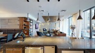 This sophisticated single-floor space showcases a thoughtfully designed open-concept layout that seamlessly blends functionality with contemporary style. The kitchen anchors the space with a striking dark granite island, stainless steel appliances, and a series of sculptural pendant lights suspended from a bright white ceiling, creating a refined focal point.

Beyond the kitchen, the open living area unfolds into distinct yet connected zones for lounging and conversation. Rich leather sofas and upholstered armchairs are arranged around a sleek coffee table, offering a warm, inviting atmosphere. A wall-mounted television is framed by a rustic wood bookshelf unit, adding texture and a residential warmth that balances the modern aesthetic.

Toward the far end of the space, a slatted divider partition provides subtle definition without interrupting the open flow, while floor-to-ceiling windows along the perimeter bathe the entire interior in abundant natural light and offer views of the surrounding greenery. The layered ceiling design, featuring recessed and dropped architectural elements with integrated lighting, adds depth and visual interest throughout.

The overall ambiance strikes a balance between polished and comfortable — a well-appointed space ideal for gathering, relaxing, or conducting business in a welcoming, design-forward environment.